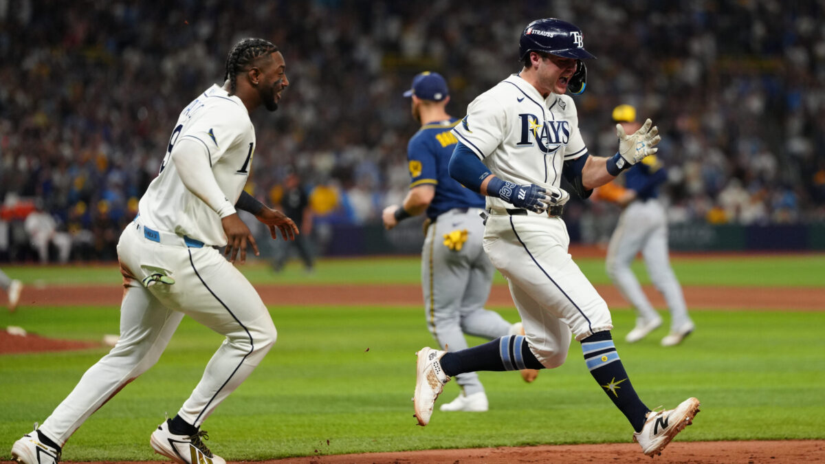 Two baseball players in white Rays uniforms celebrate on the field as opponents walk in the background.