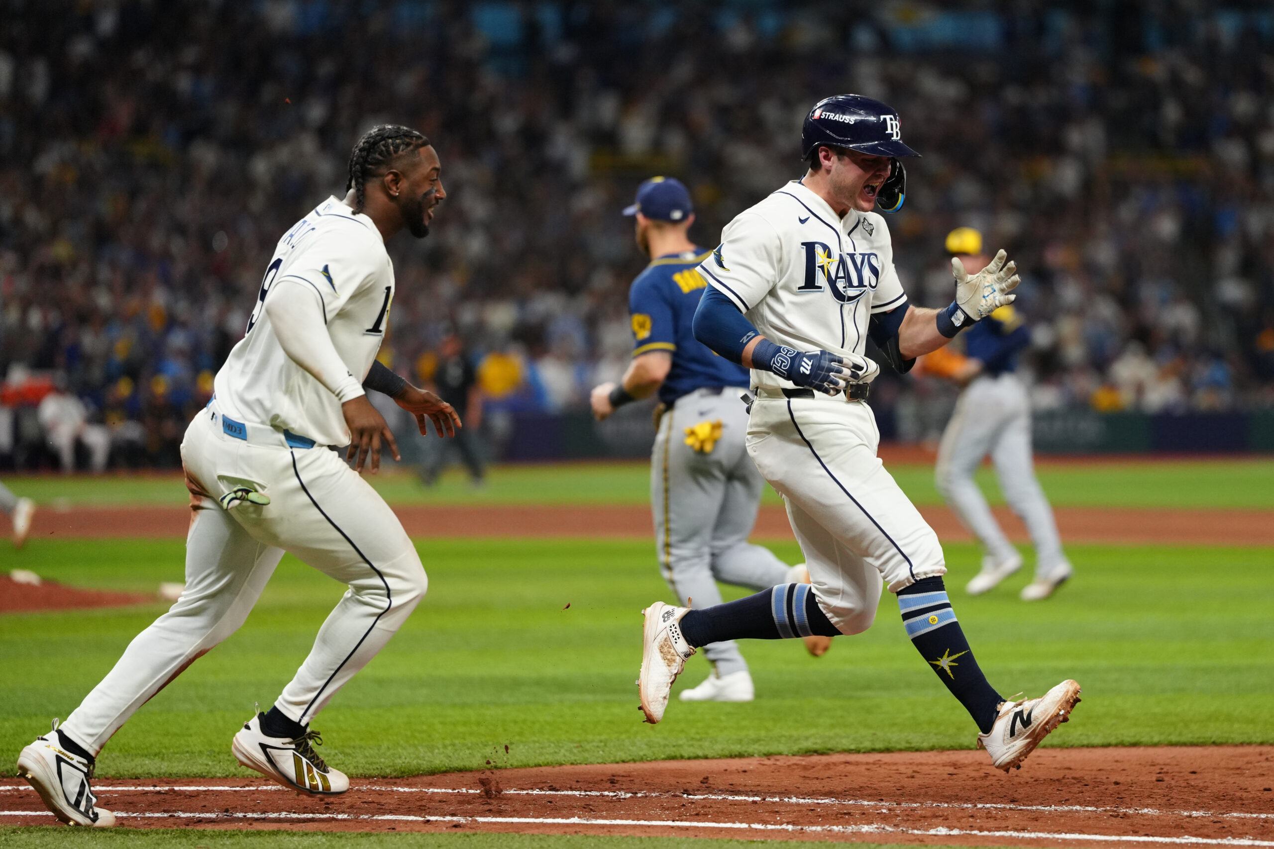 Two baseball players in white Rays uniforms celebrate on the field as opponents walk in the background.