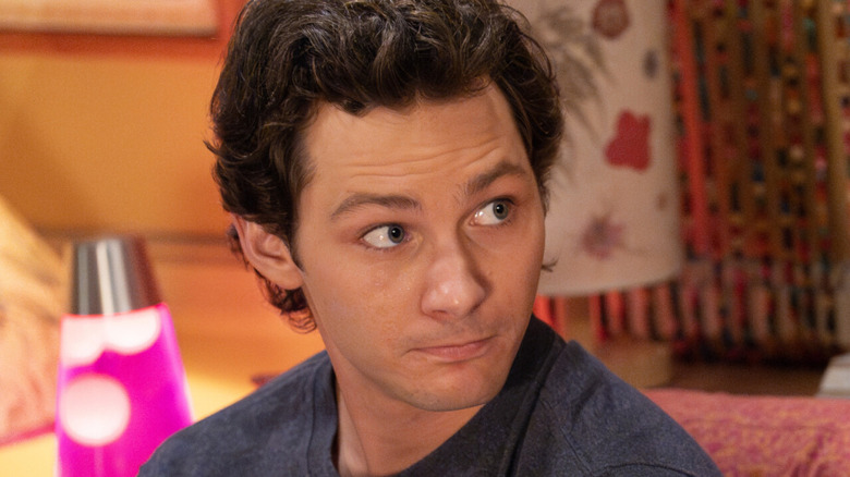 A young man with curly hair looks to the side beside a glowing pink lamp in a warmly lit room.