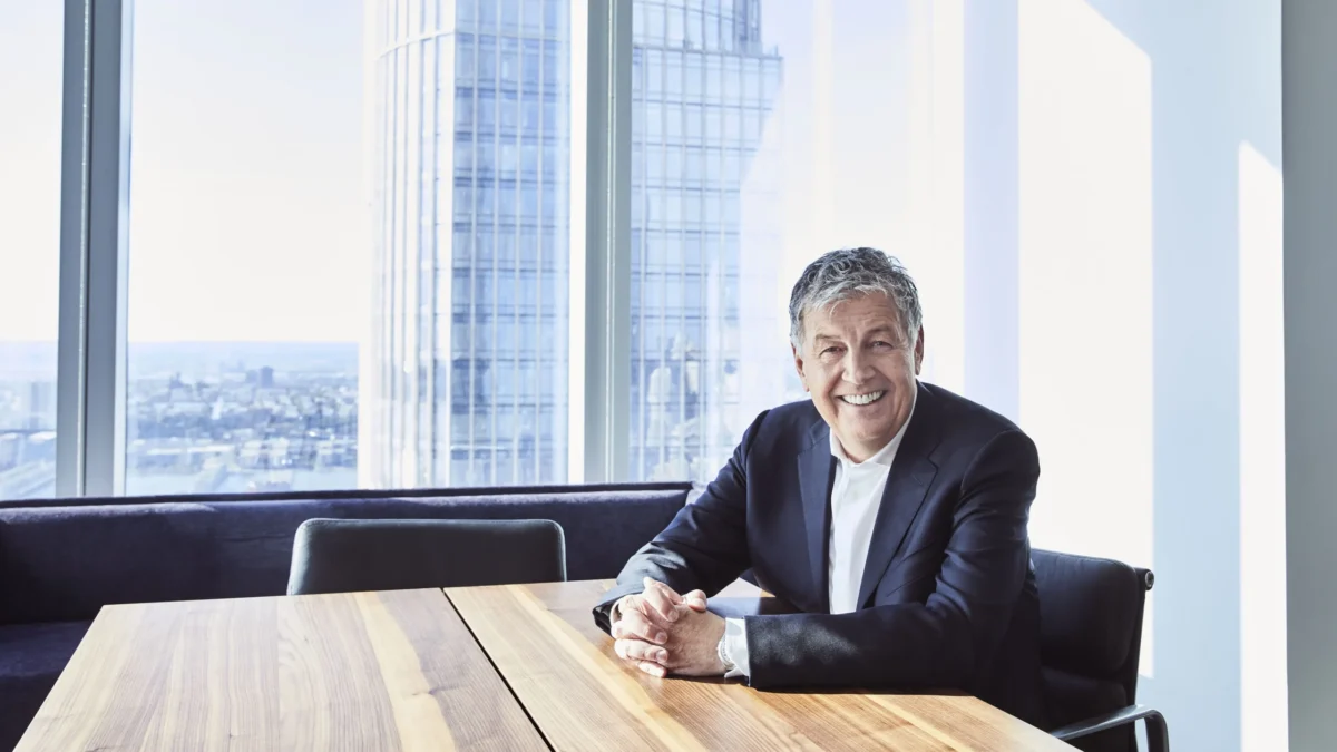 Smiling gray-haired man in a dark suit sits at a wooden table beside large windows overlooking city buildings.