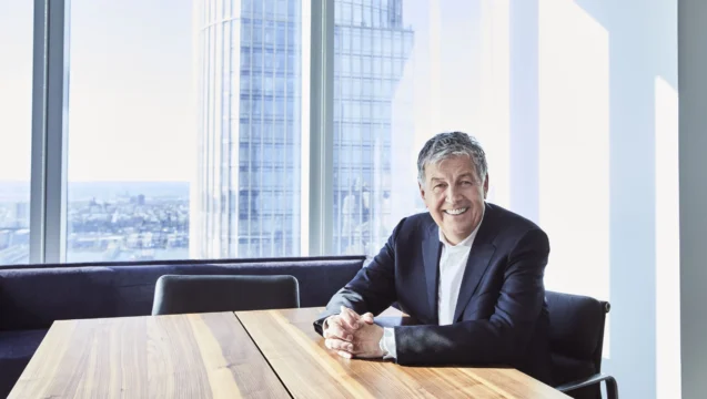 Smiling gray-haired man in a dark suit sits at a wooden table beside large windows overlooking city buildings.
