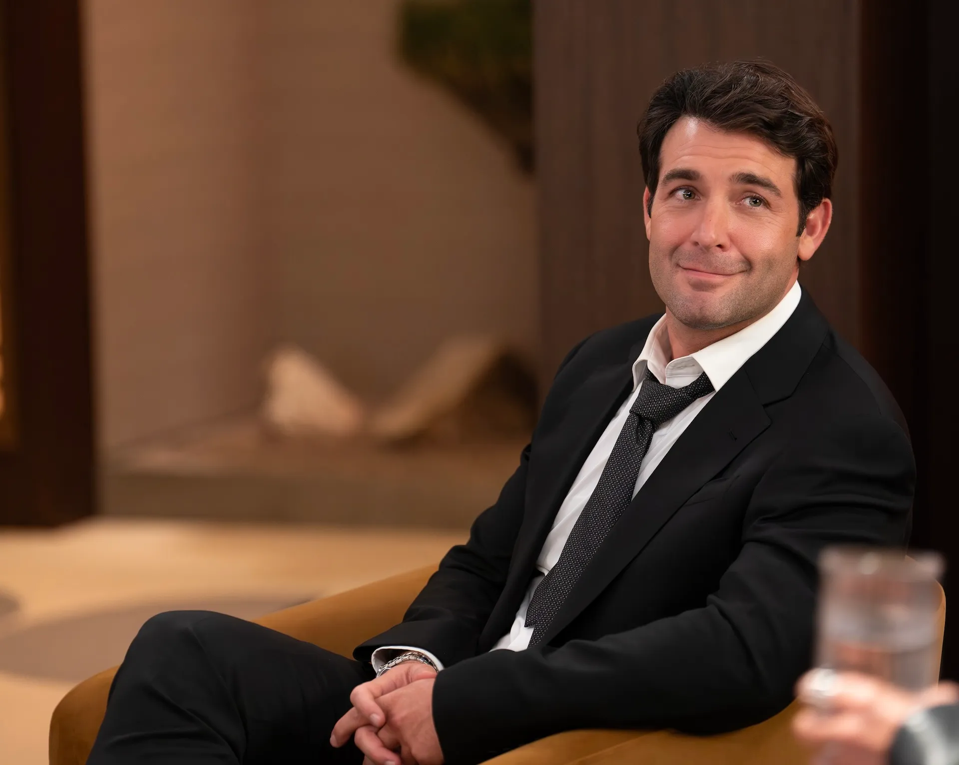 A man in a black suit and loosened tie sits in a chair, smiling slightly.