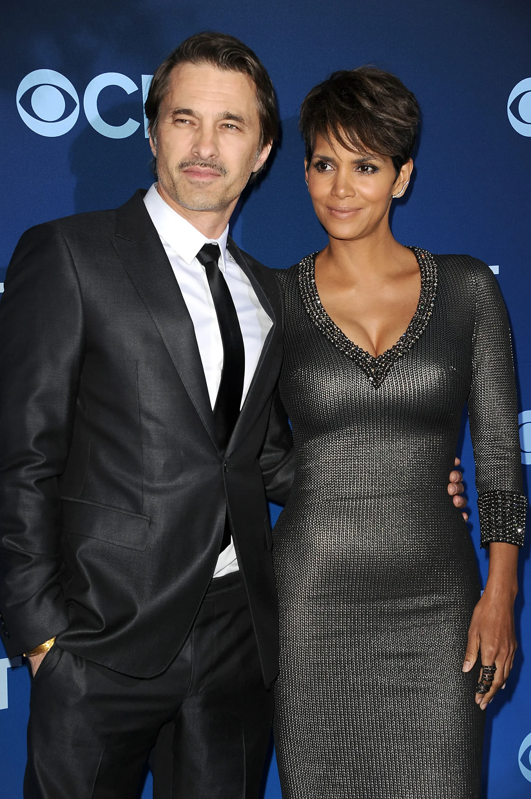 Two formally dressed people pose together in front of a blue backdrop with white logos.