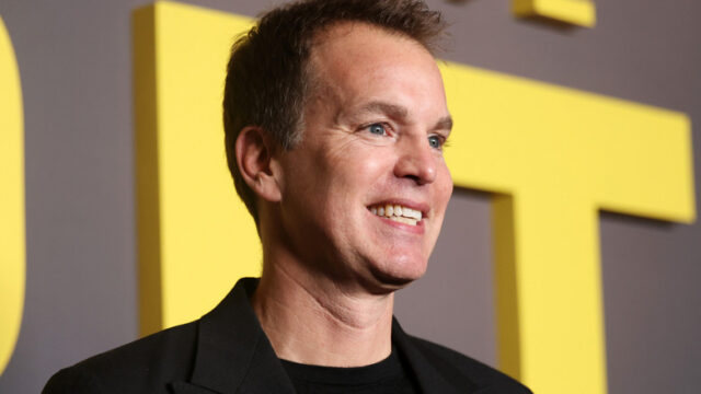 A smiling man in a black jacket stands before large yellow letters on a gray backdrop.