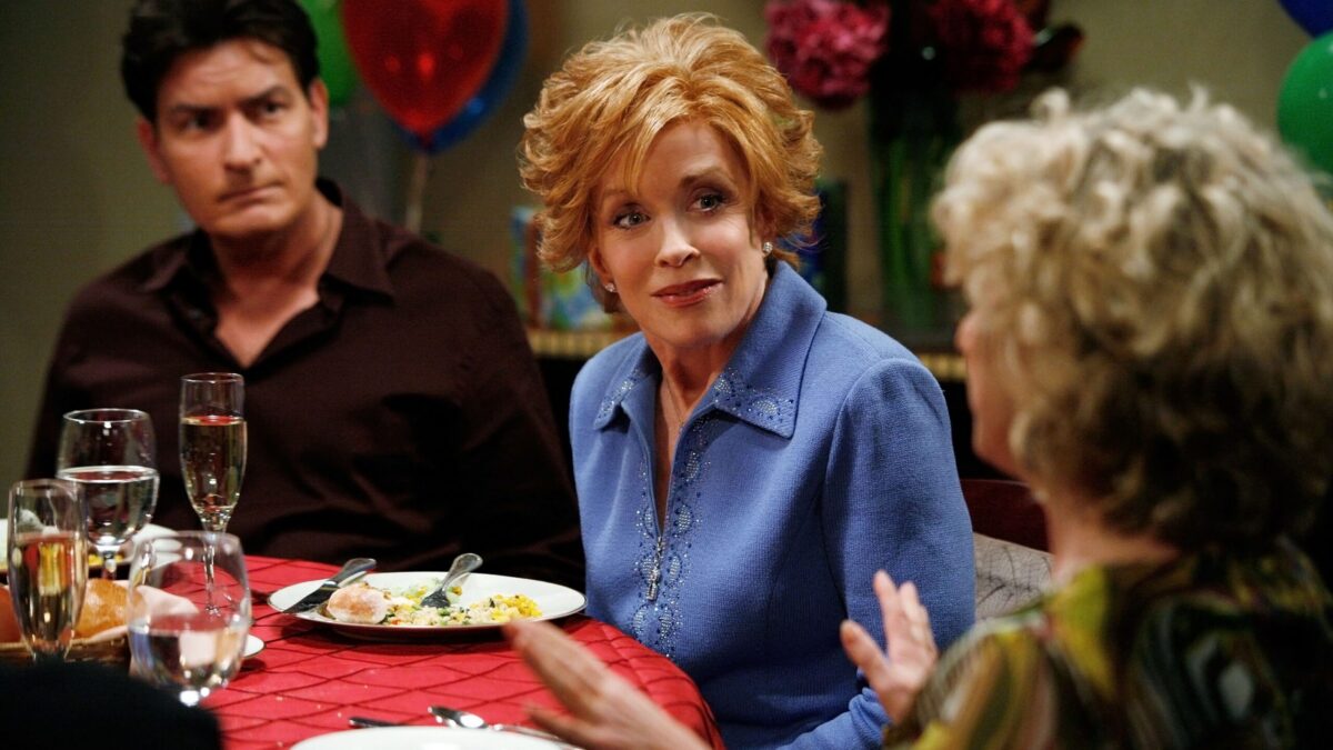 A woman in a blue sweater sits at a dinner table with others, balloons and flowers behind them.