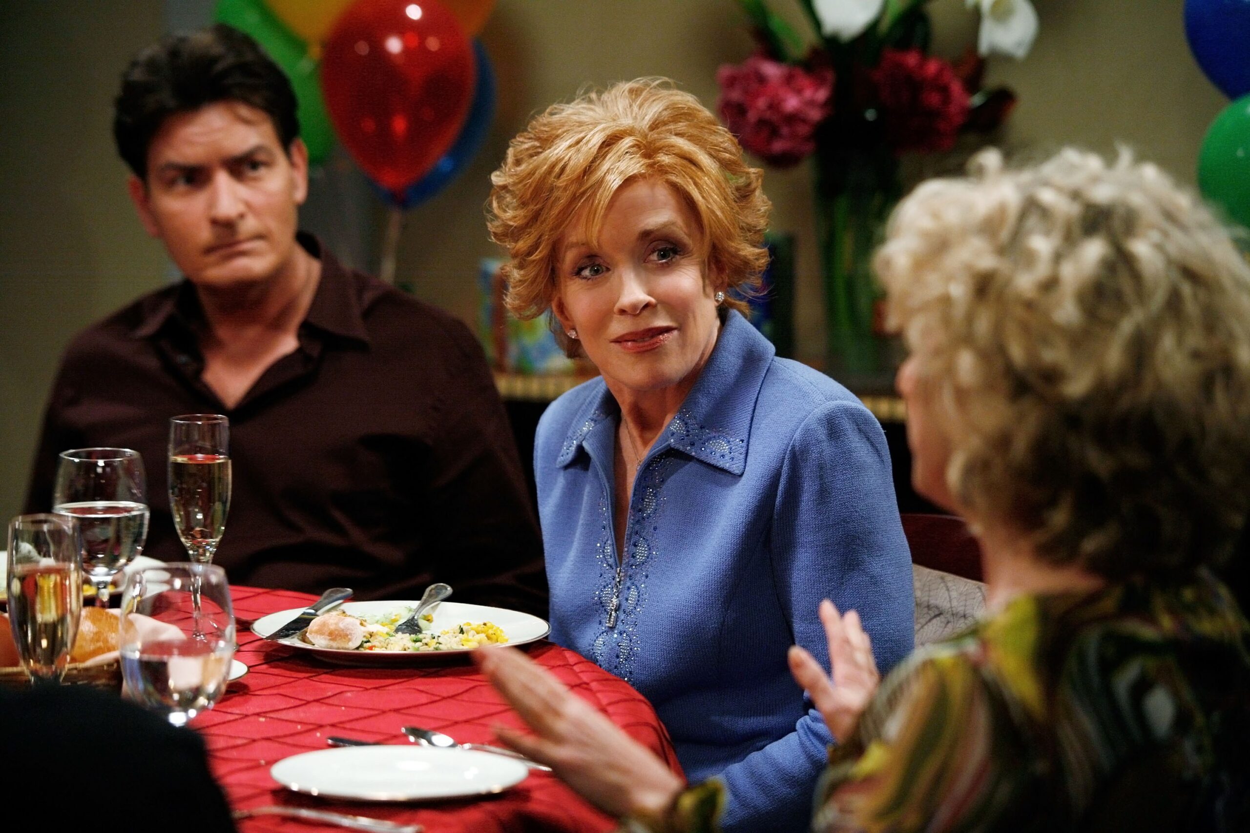 A woman in a blue sweater sits at a dinner table with others, balloons and flowers behind them.