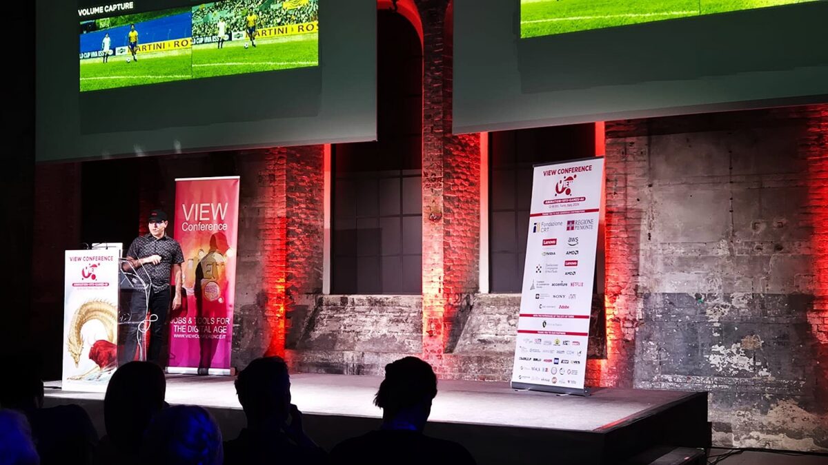 A person stands on a conference stage beneath large screens showing soccer footage, with audience silhouettes in front.