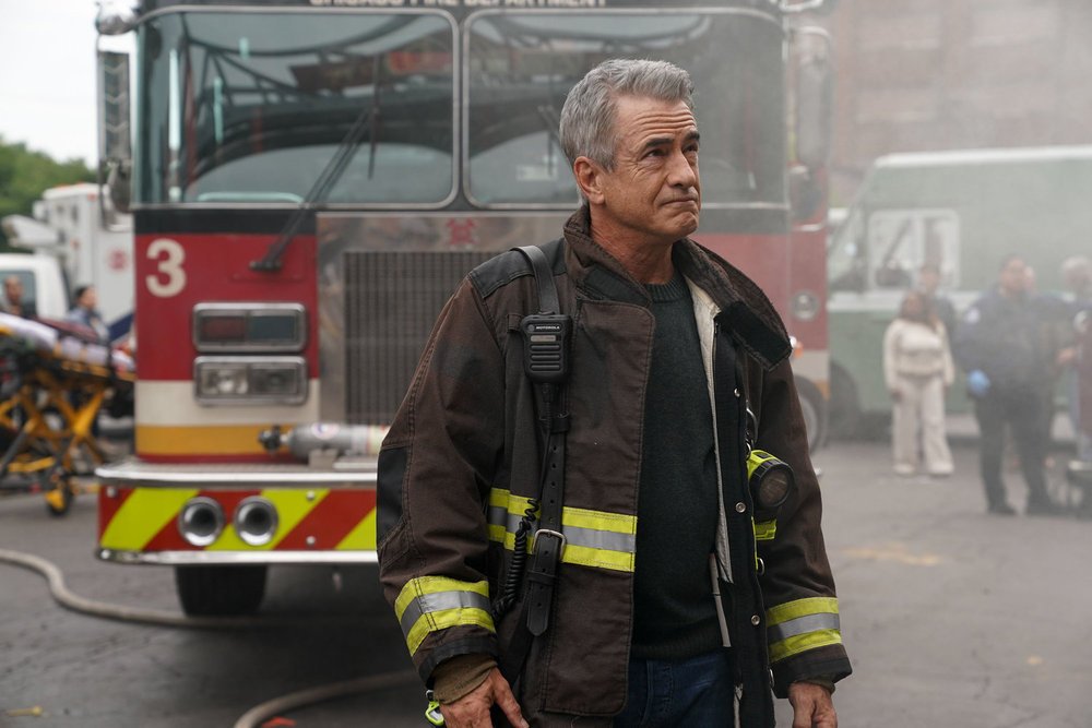 A gray-haired firefighter in turnout gear stands before a fire truck amid smoky emergency activity.