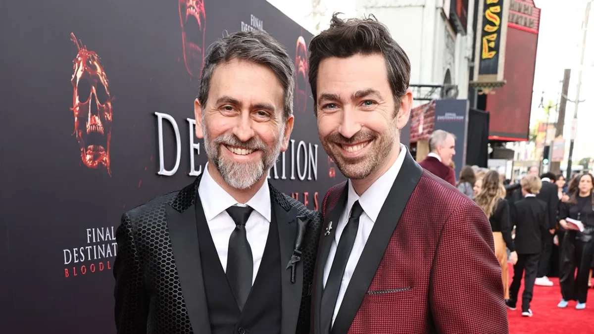 Two smiling men in suits pose on a red carpet beside a dark movie poster backdrop.