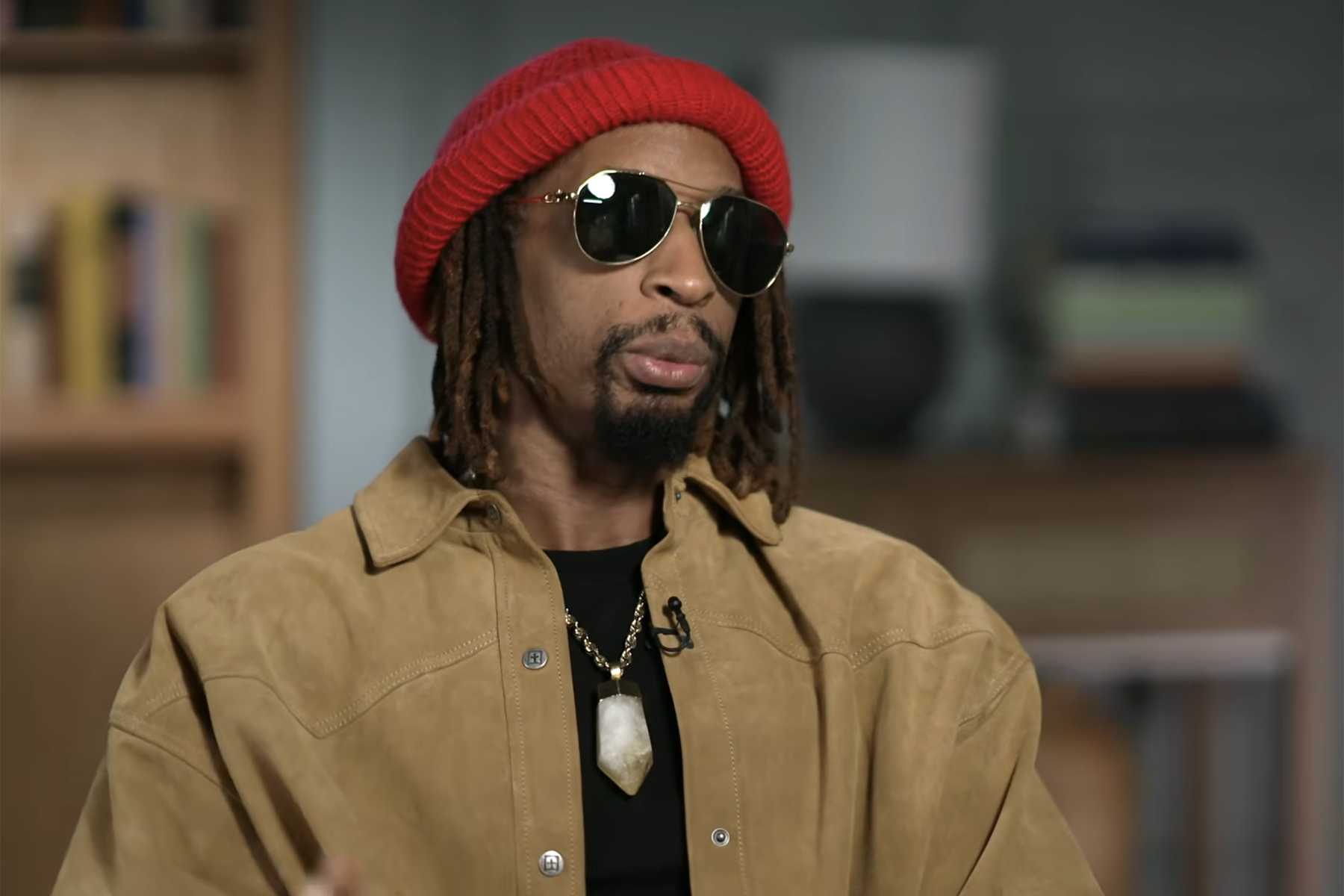 A man in a red knit hat and sunglasses wears a tan jacket against a blurred indoor background.