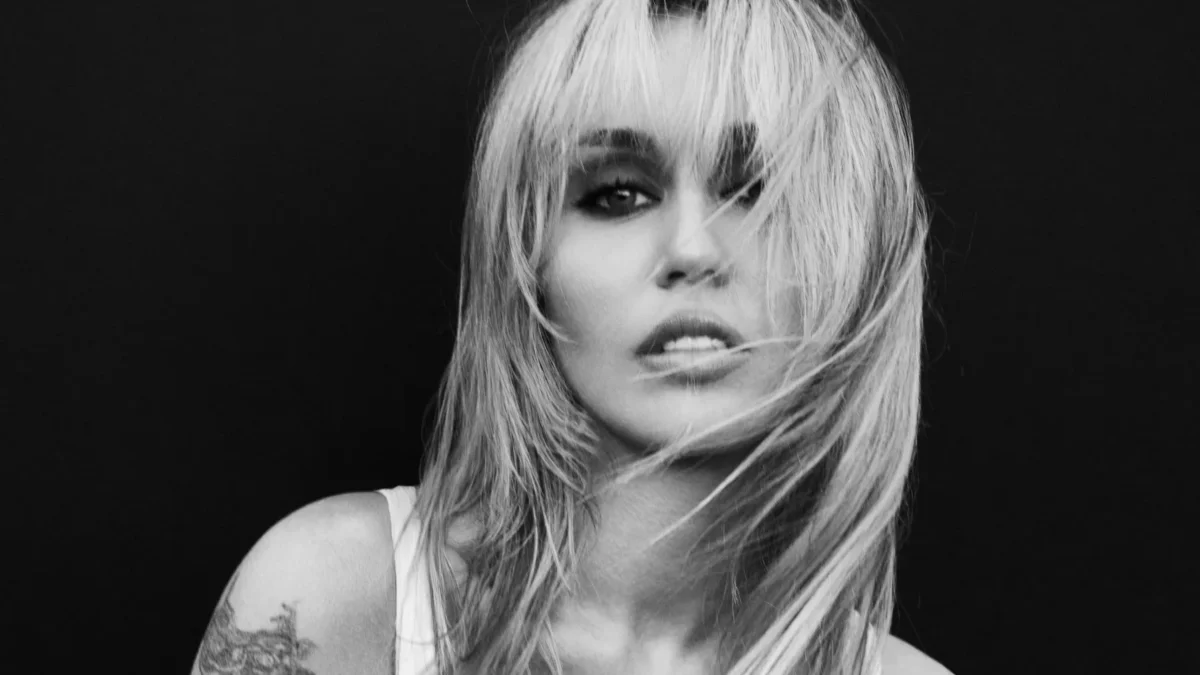 Black-and-white close-up of a woman with tousled light hair, dark eye makeup, and a shoulder tattoo.