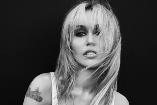 Black-and-white close-up of a woman with tousled light hair, dark eye makeup, and a shoulder tattoo.