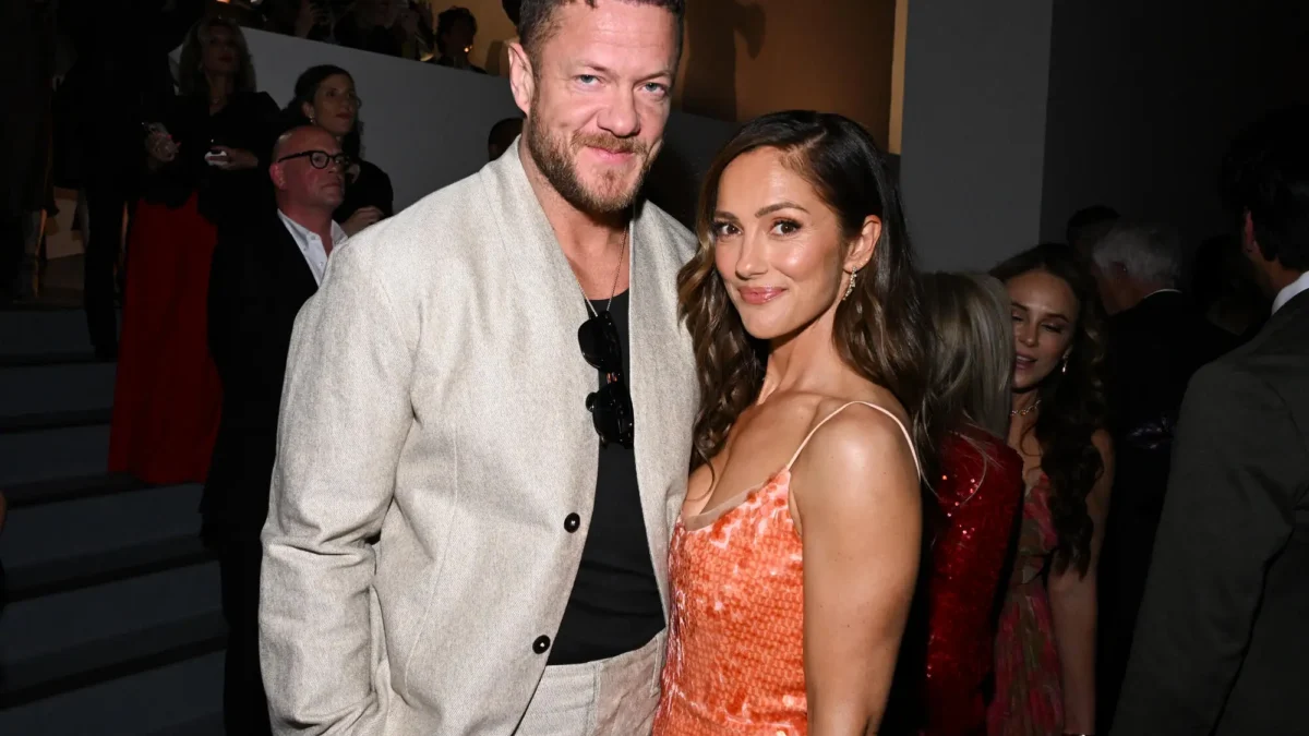 A man in a light suit and a woman in an orange dress pose together at a crowded indoor event.