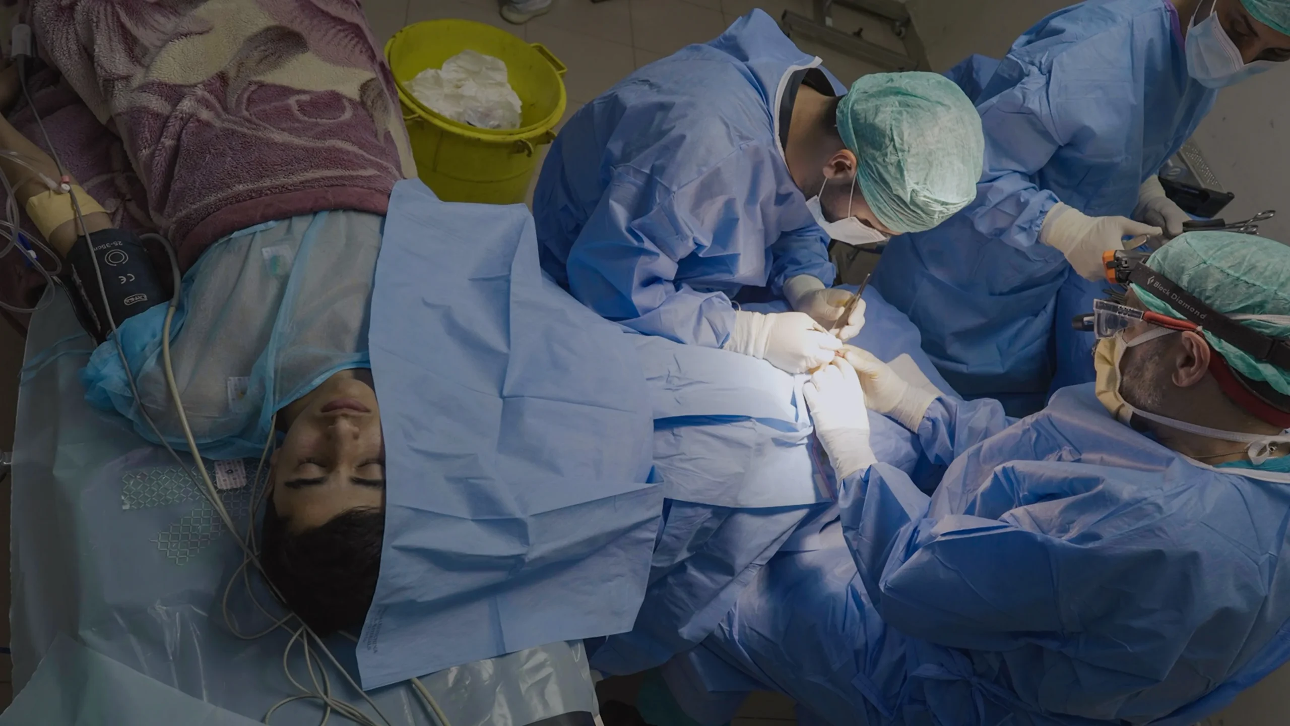 Surgeons in blue gowns operate on a draped patient lying under monitors and blankets.