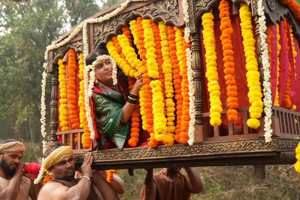 A woman in green attire sits in a flower-draped palanquin carried by several men outdoors.