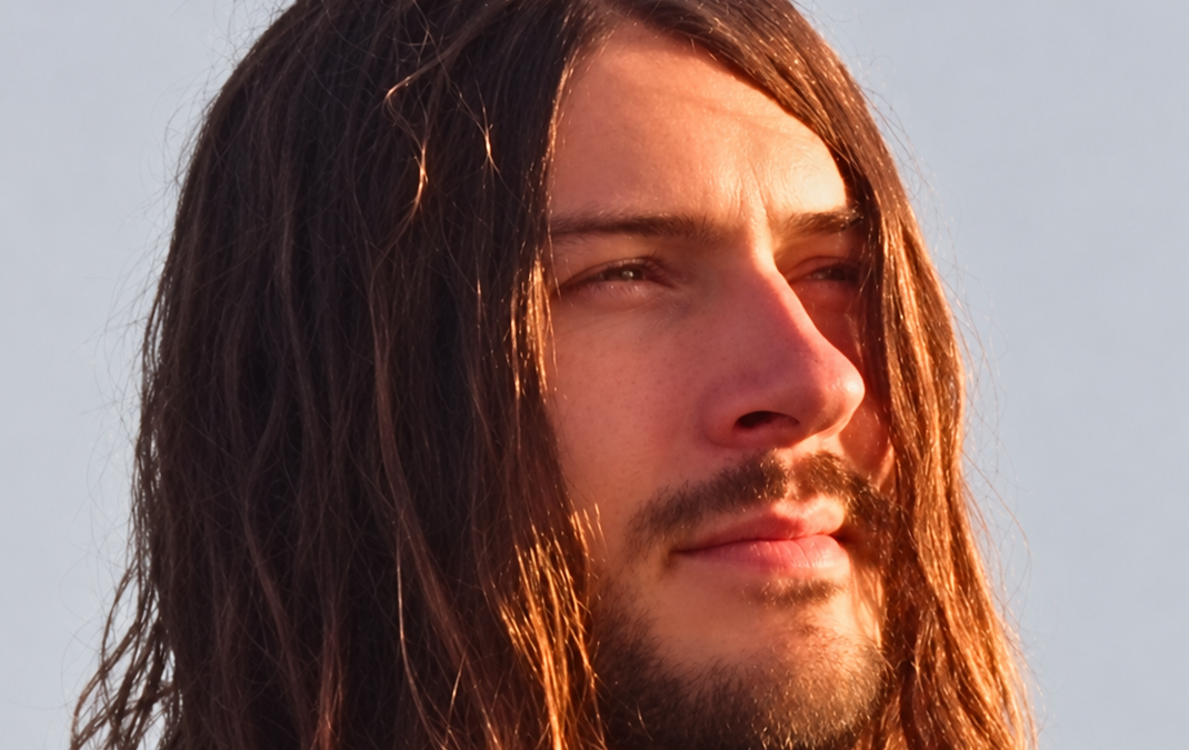 Close-up of a long-haired man with a beard looking into warm sunlight against a pale sky.