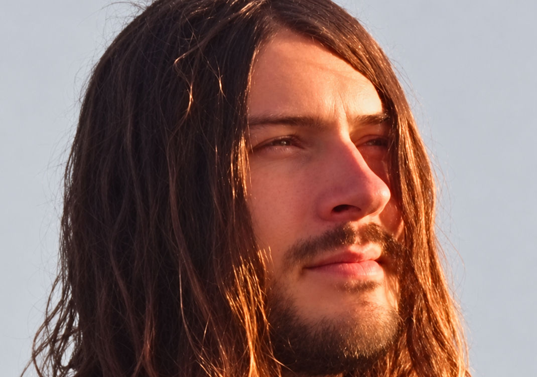Close-up of a long-haired man with a beard looking into warm sunlight against a pale sky.