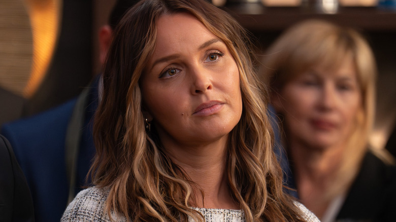A woman with wavy hair looks thoughtful while another woman sits blurred in the background.