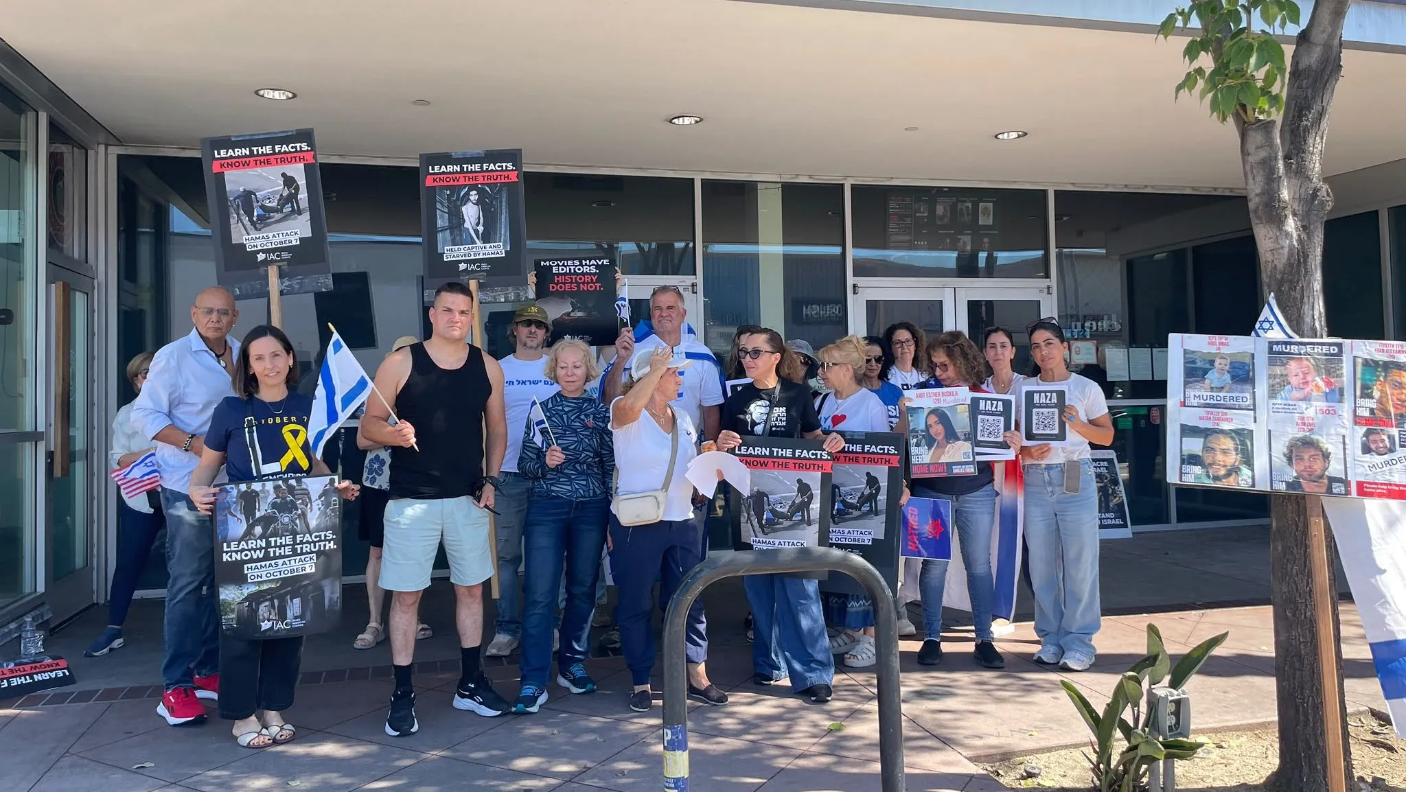 A group stands outside a building holding signs, posters, and blue-and-white flags.