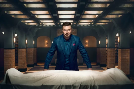 A man in a blue suit leans over a white sheet-covered form in a dim tiled room.