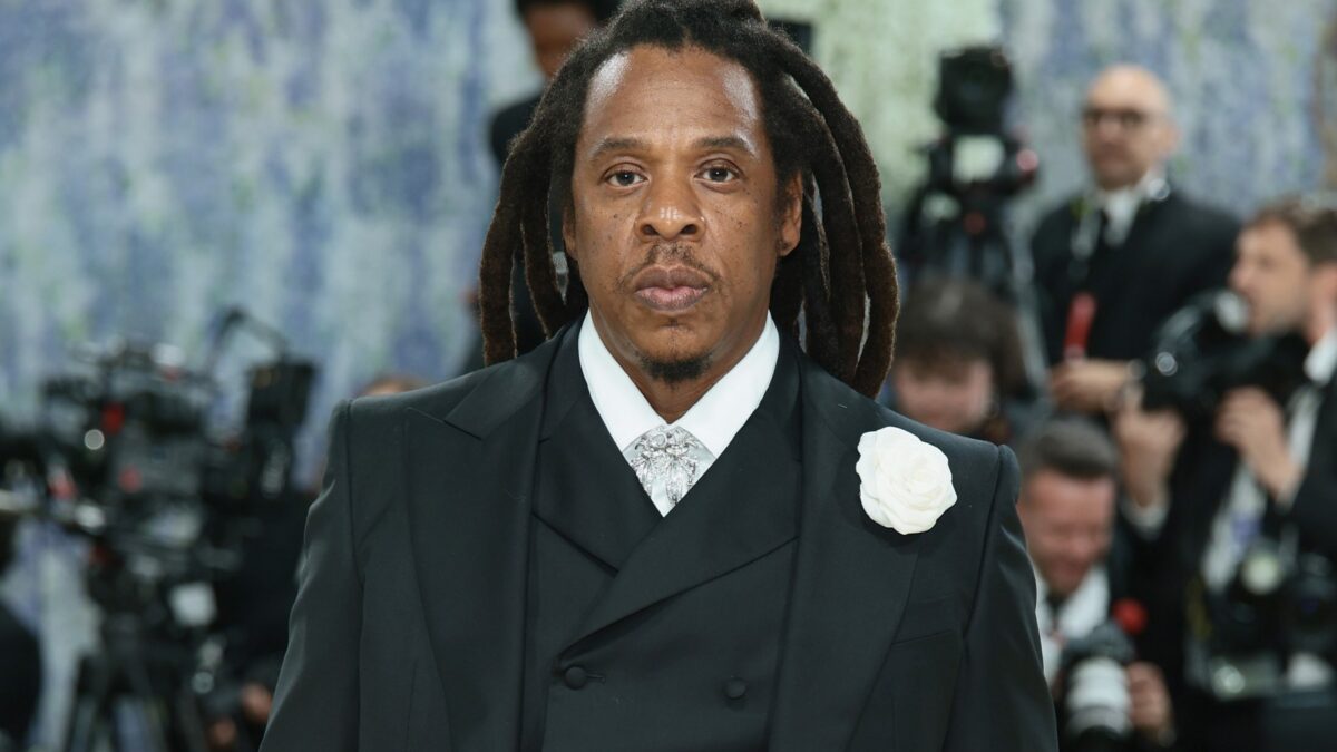 A man with long locs in a black suit and white rose boutonniere stands before photographers.