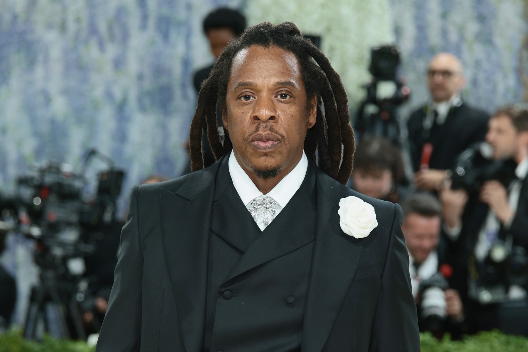 A man with long locs in a black suit and white rose boutonniere stands before photographers.