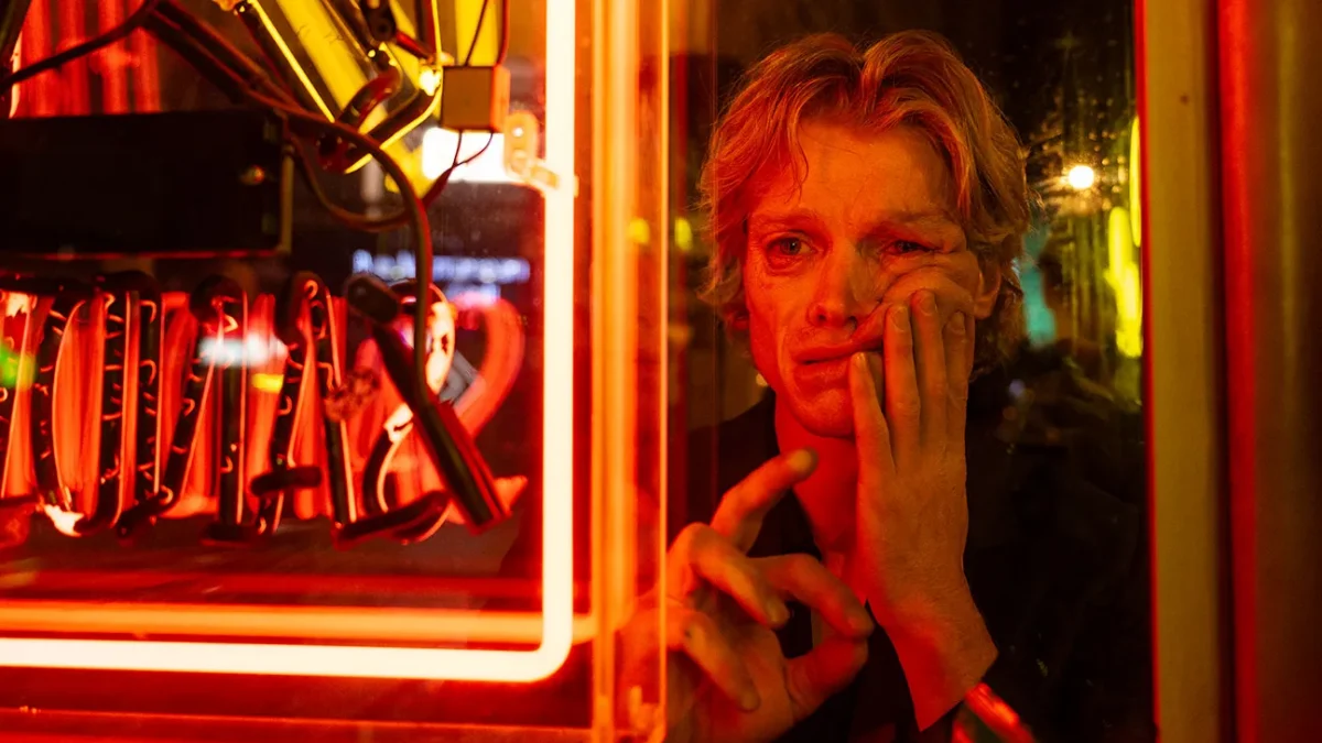 A man presses his face and hand against a window beside glowing red neon signs.