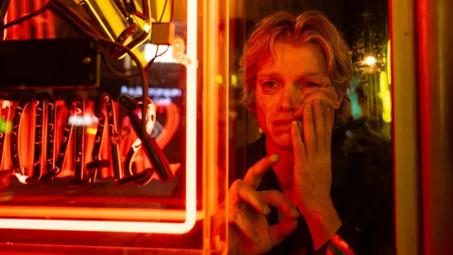 A man presses his face and hand against a window beside glowing red neon signs.