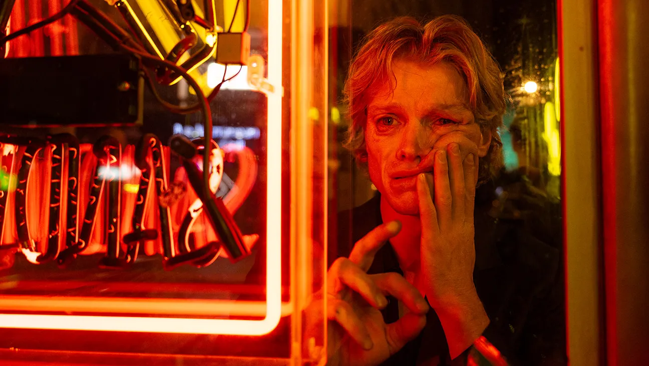 A man presses his face and hand against a window beside glowing red neon signs.