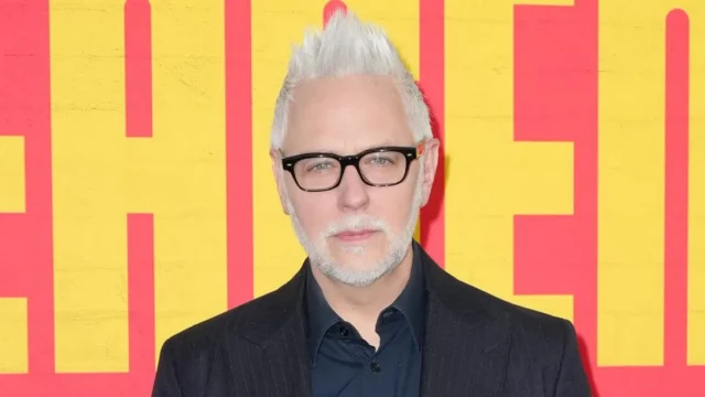 Man with spiked white hair and black glasses poses in front of a yellow and red backdrop.