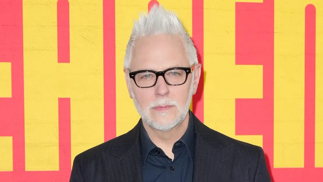 Man with spiked white hair and black glasses poses in front of a yellow and red backdrop.