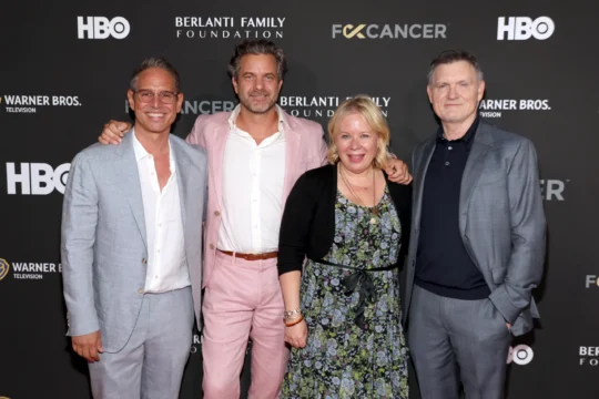 Four people pose together on a black logo backdrop, wearing suits and a floral dress.