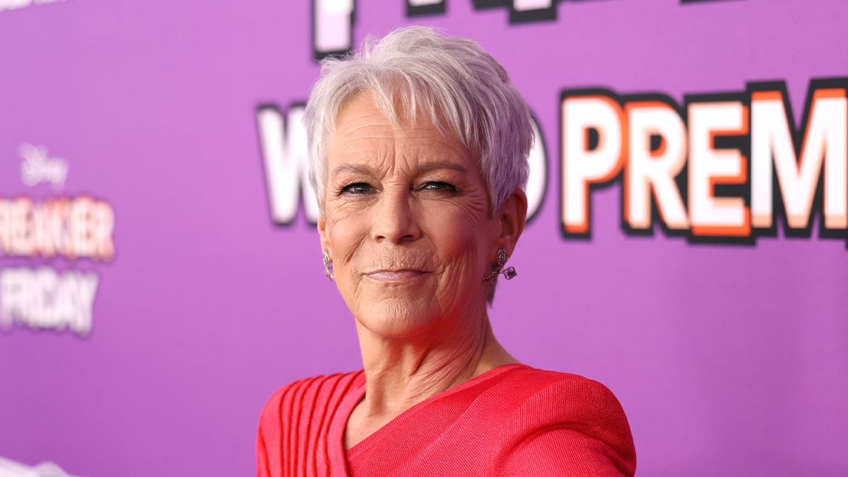 A woman with short gray hair wears a bright red dress against a purple backdrop.