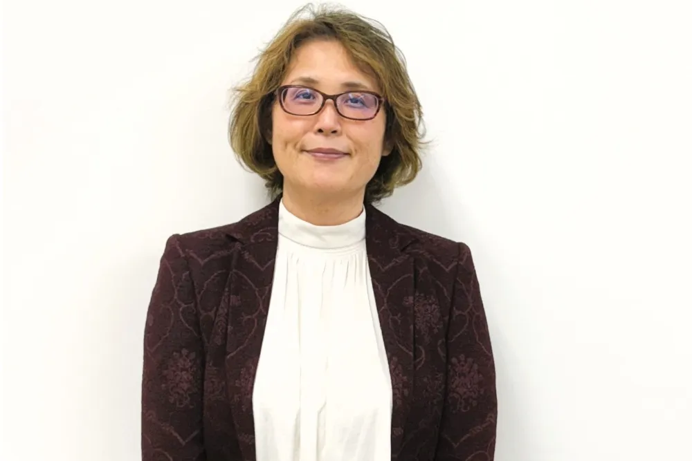 A person with glasses and short hair wears a dark patterned blazer against a plain light background.
