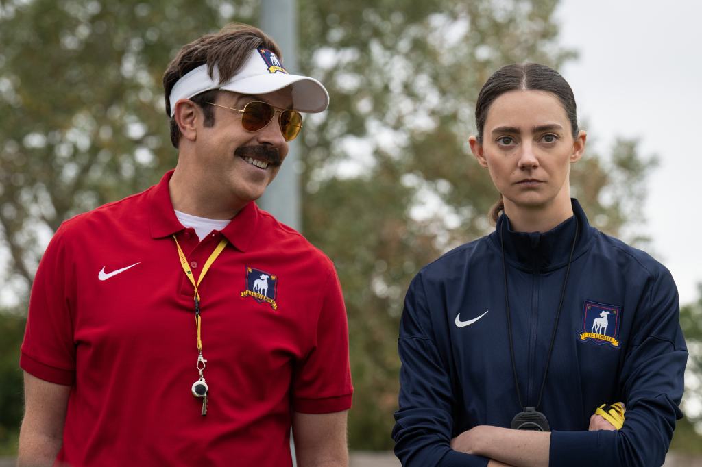 A man in a red polo smiles beside a serious woman in a navy jacket, both wearing whistles.