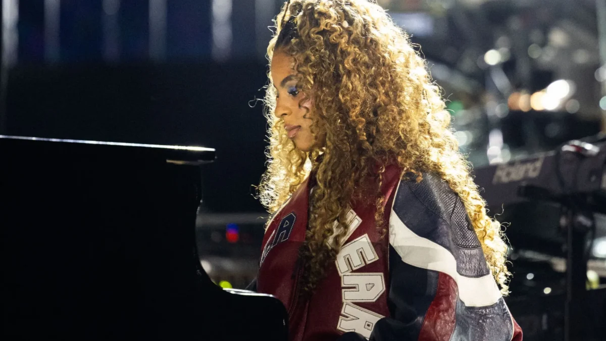 A curly-haired person in a colorful jacket sits at a piano under bright stage lights.