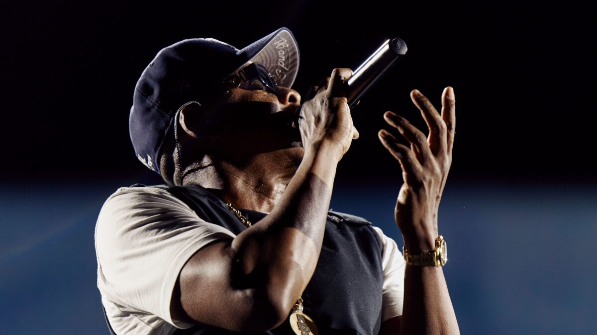 A man in a cap and sunglasses holds a microphone and raises one hand against a dark background.