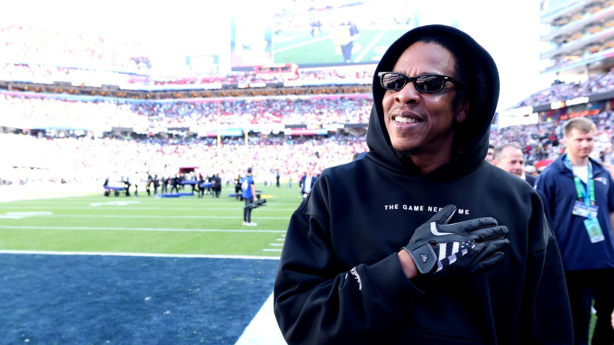 Man in black hoodie and sunglasses stands on a football field with hand on chest in a crowded stadium.