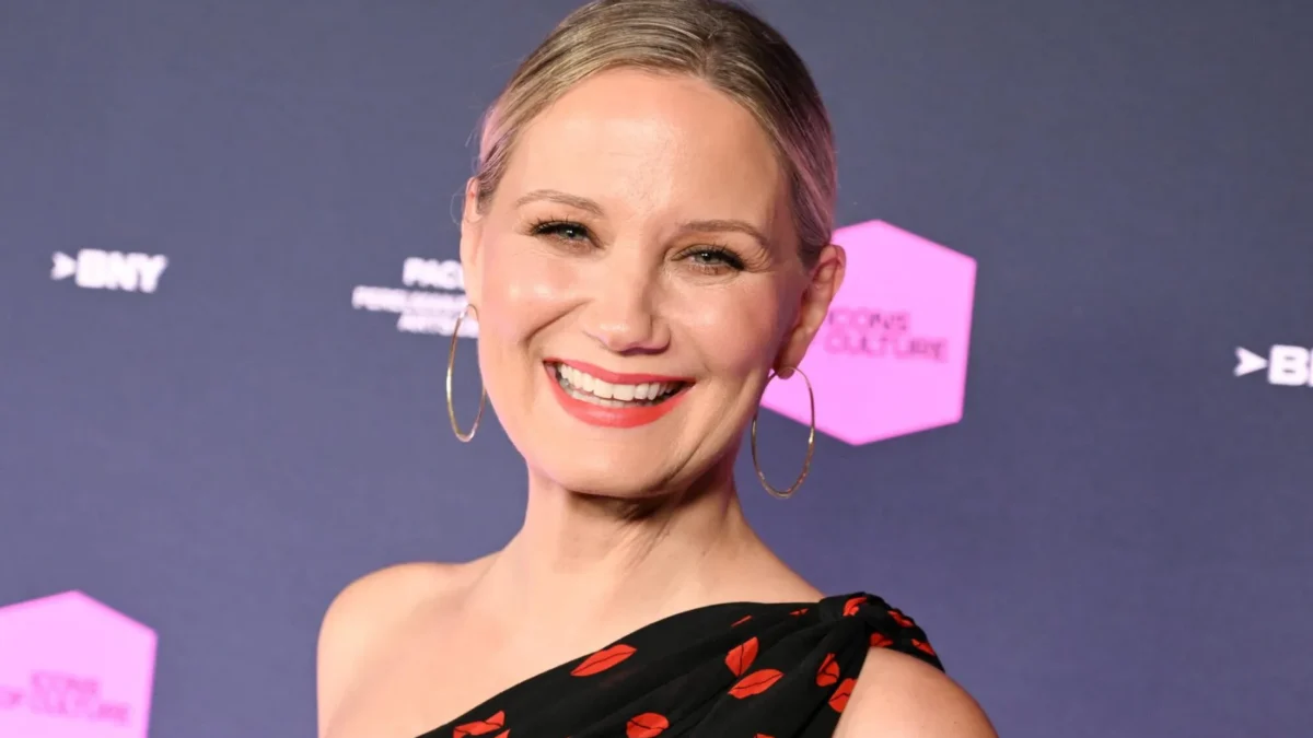 Smiling blonde woman in hoop earrings and a black one-shoulder dress with red lip prints against a dark backdrop.