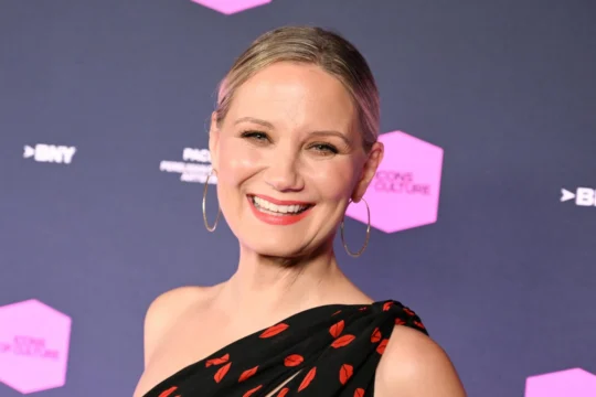 Smiling blonde woman in hoop earrings and a black one-shoulder dress with red lip prints against a dark backdrop.