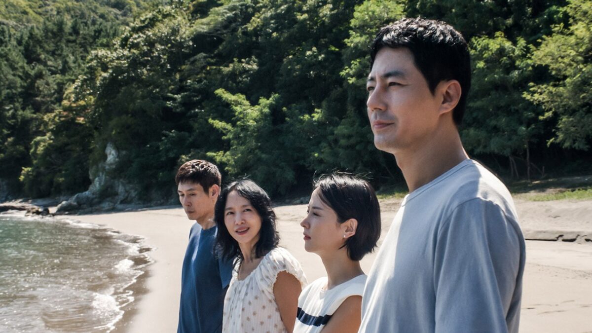 Four people stand on a sandy beach beside calm waves, with dense green trees and rocks behind them.