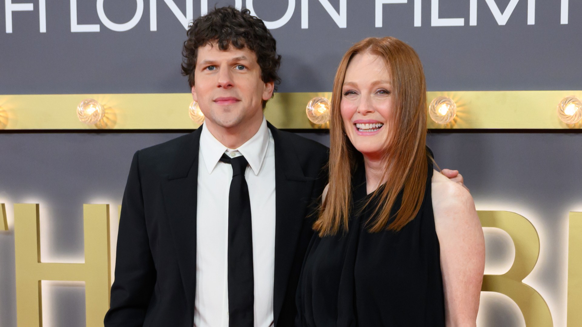 Two people dressed in black pose together before a lit gray-and-gold backdrop.