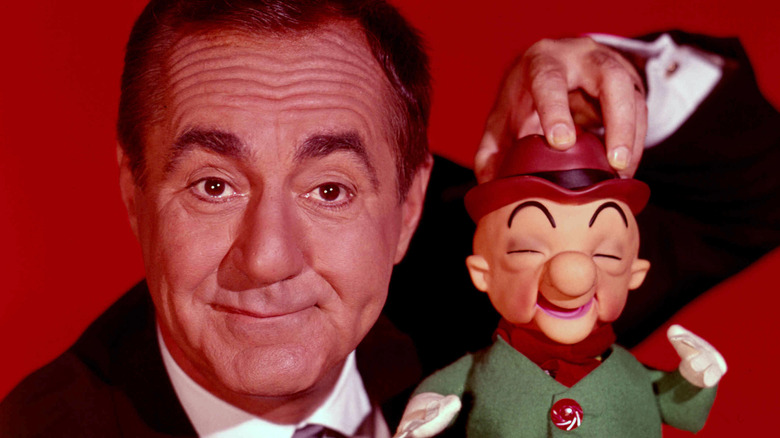 A man in a tuxedo poses beside a smiling puppet in a hat against a red background.