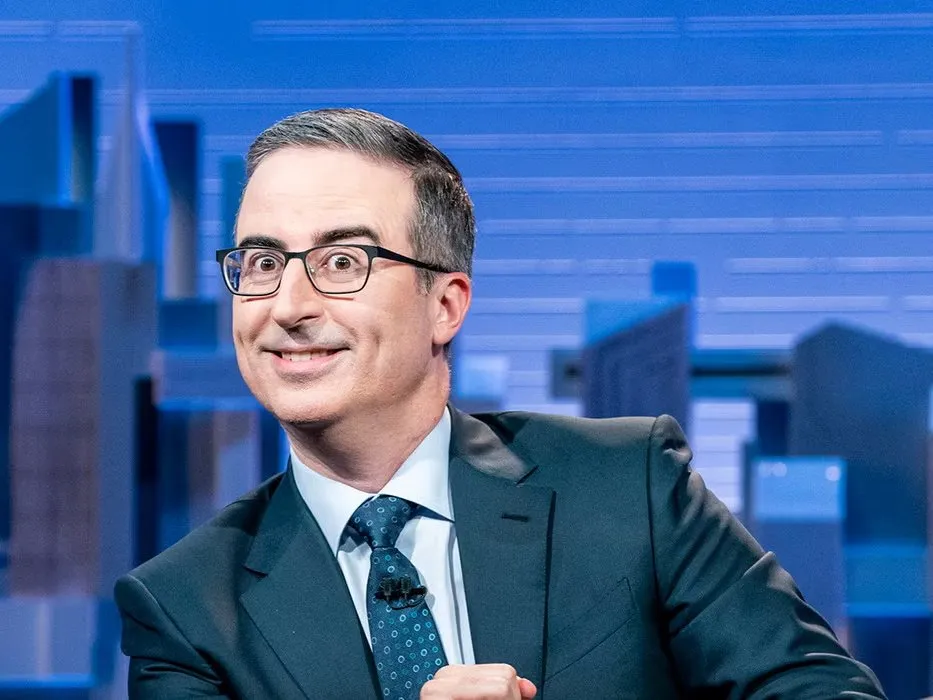A man in glasses and a suit smiles in front of a blue studio backdrop.