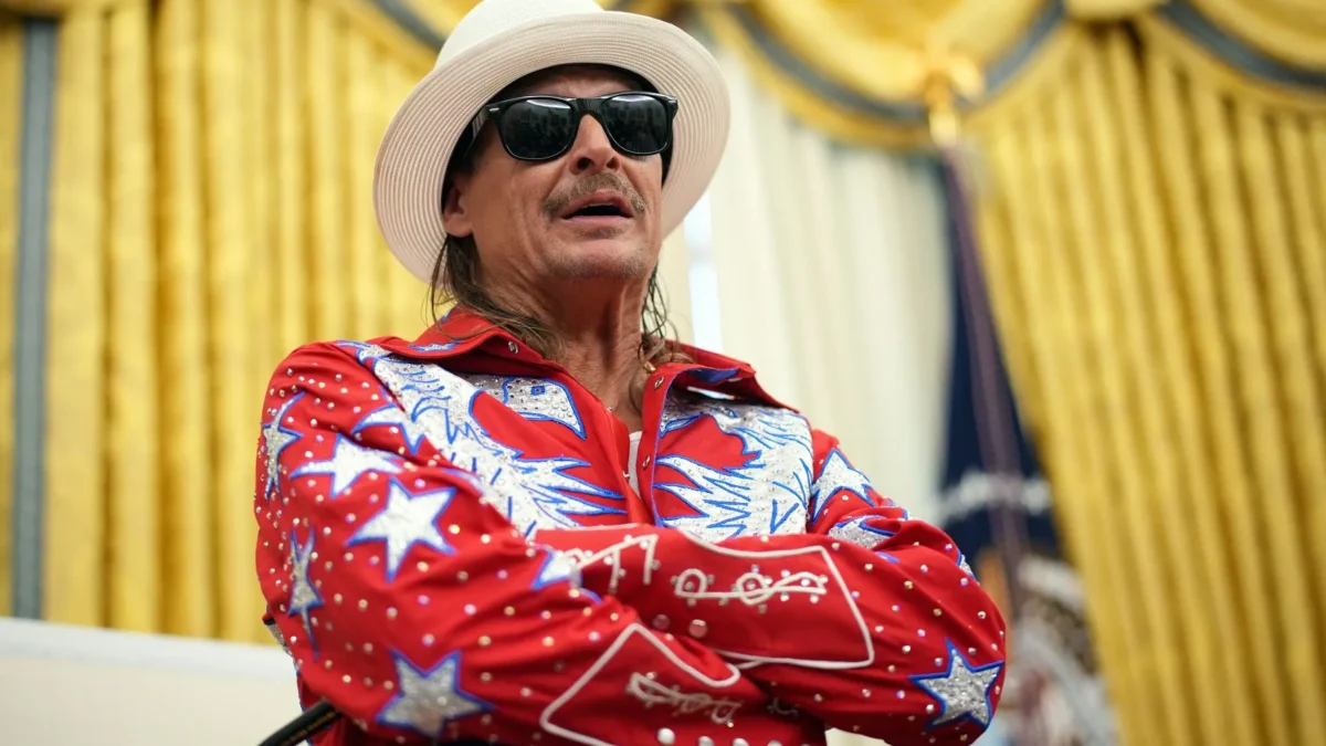 A man in sunglasses and a white hat wears a red sequined star-patterned outfit before gold curtains.