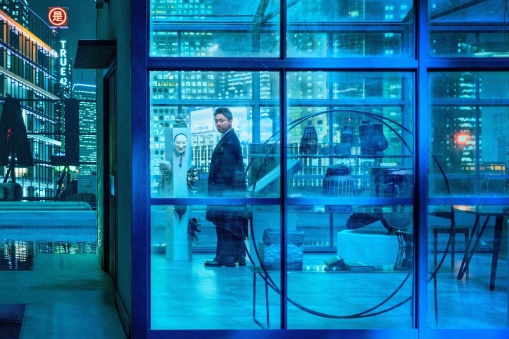 A man in a suit stands beside a mannequin inside a blue-lit glass room overlooking city lights.