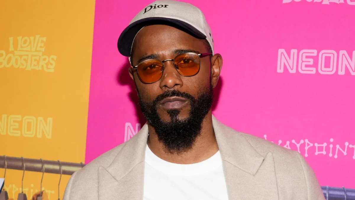 A bearded man wearing tinted glasses and a beige cap poses before a bright pink and orange backdrop.