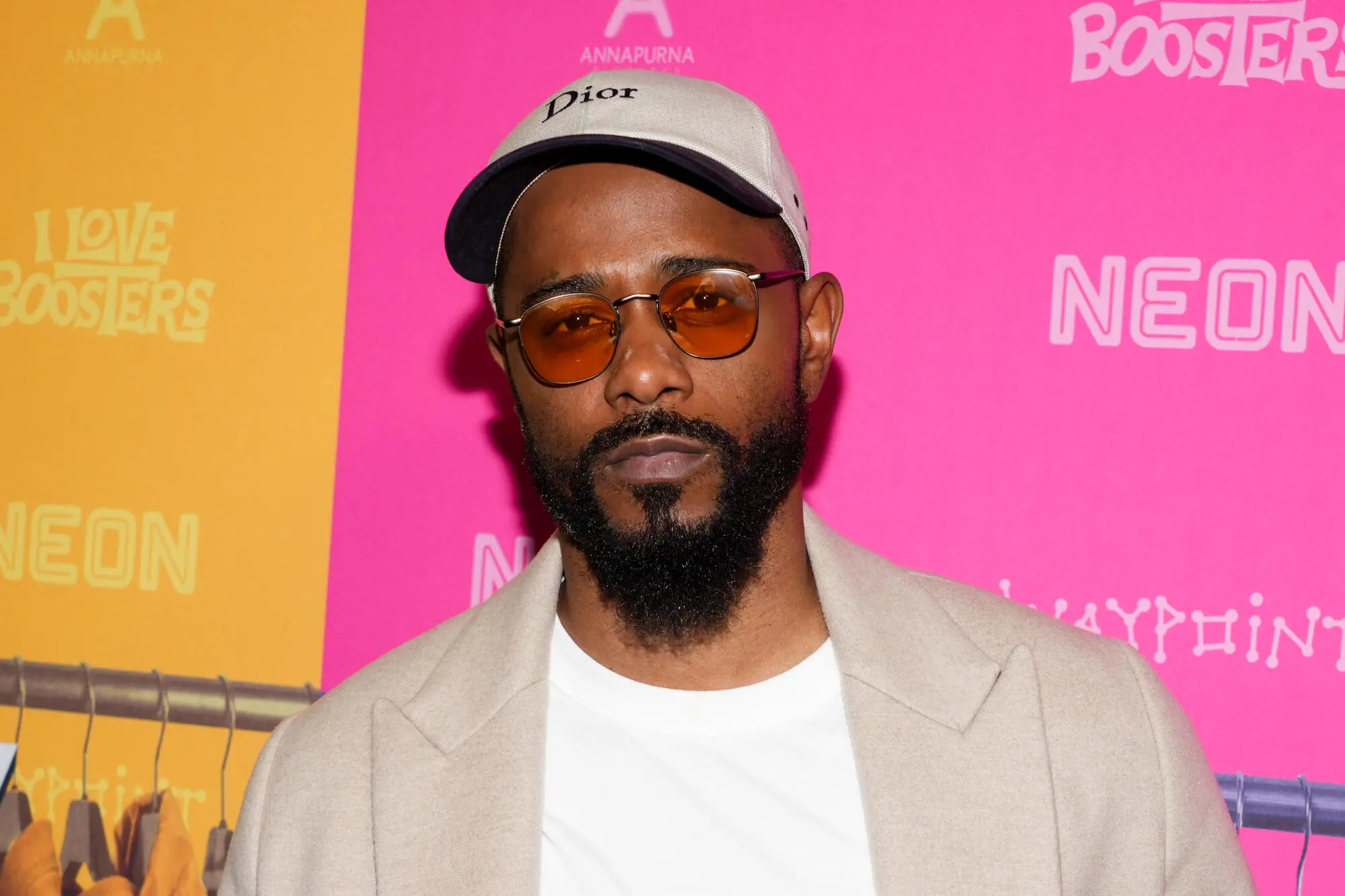 A bearded man wearing tinted glasses and a beige cap poses before a bright pink and orange backdrop.