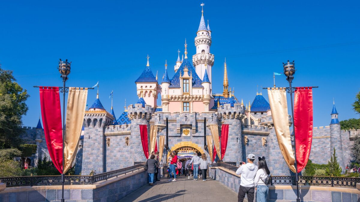A fairy-tale castle with blue spires rises behind red and gold banners as visitors walk across a bridge.