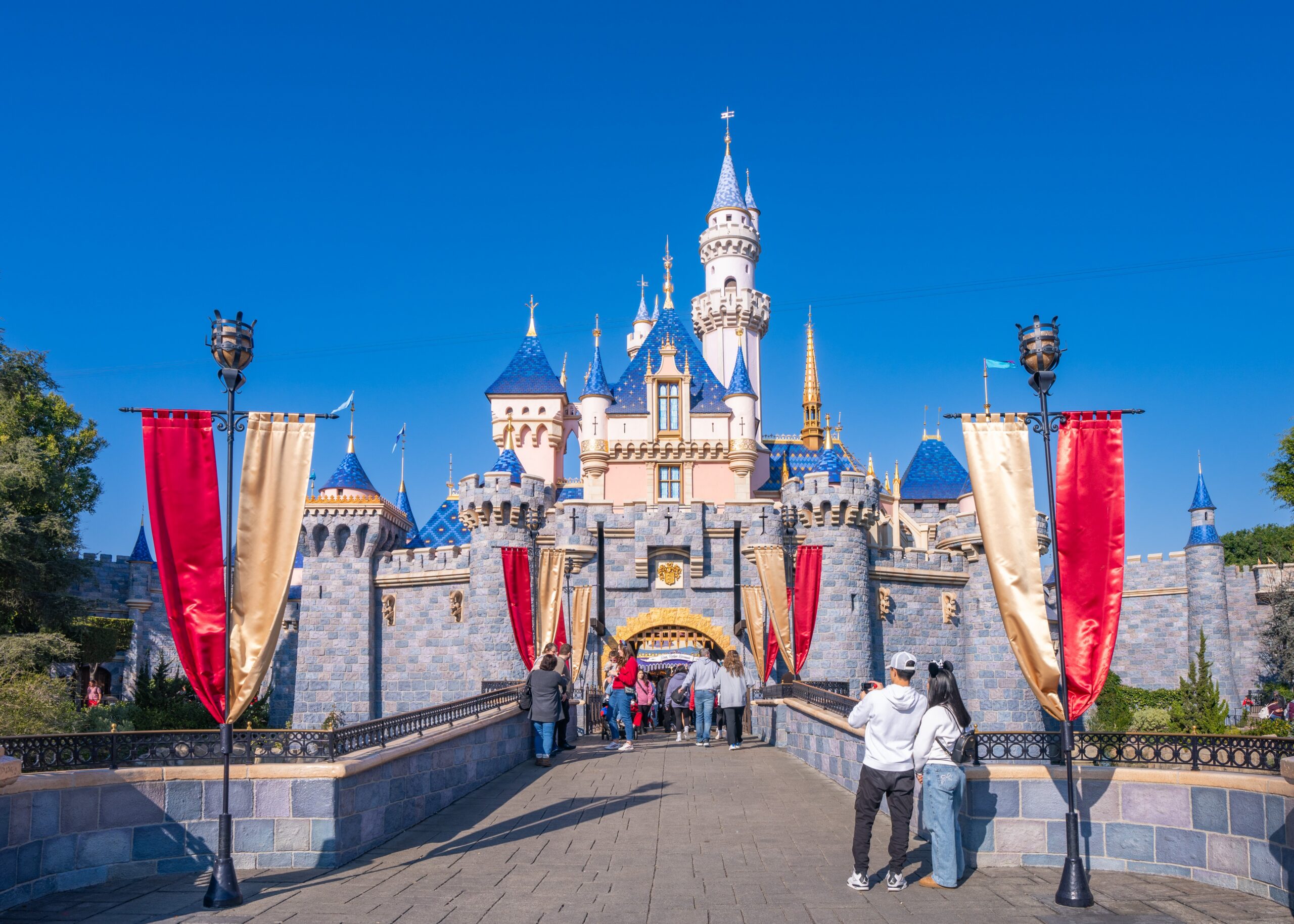 A fairy-tale castle with blue spires rises behind red and gold banners as visitors walk across a bridge.